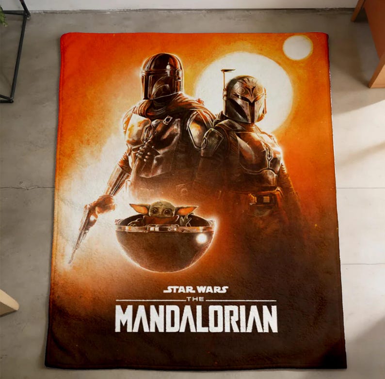 K&ouml;nnte beinhalten: Eine rechteckige Decke mit Figuren aus "Star Wars: The Mandalorian". Das Design zeigt zwei Figuren in R&uuml;stungen, eine kleine Kreatur in einer Kapsel und den Titel der Show. Der Hintergrund ist ein Farbverlauf aus Orange und Braun.