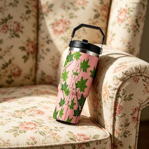 Puede incluir: Un vaso rosa con asa y tapa negra, con hiedra verde y las letras blancas "AKA". El vaso está colocado sobre un sillón con estampado floral.