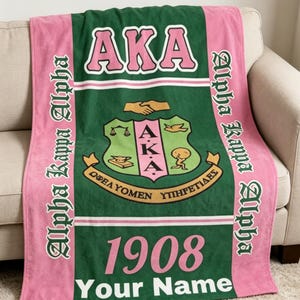 Op de afbeelding: Een groene en roze deken met de letters "AKA" in roze. De deken heeft een wapen, het jaar "1908" en de woorden "Your Name". De woorden "Alpha Kappa Alpha" staan verticaal op de roze randen.