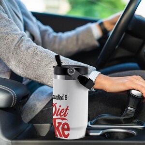 May include: White insulated tumbler with a black lid and handle. The tumbler has the words "Exhausted & Diet Coke" printed in black and red. The tumbler is in a car.