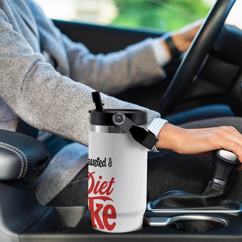 May include: White insulated tumbler with a black lid and handle. The tumbler has the words "Exhausted & Diet Coke" printed in black and red. The tumbler is in a car.