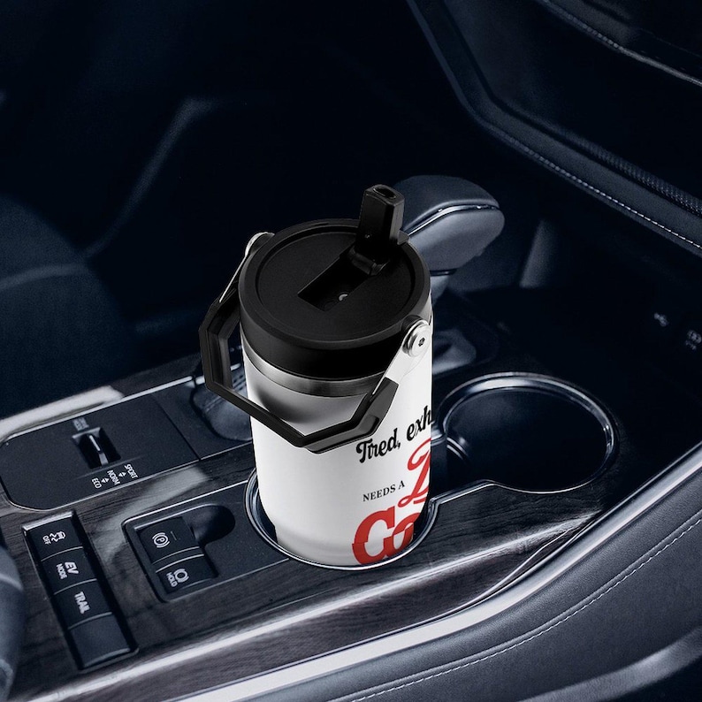 May include: A white and silver travel mug with a black lid and handle, resting in a car's cup holder. The mug has the text "Tired, exhausted, NEEDS A COFFEE" in black and red lettering.