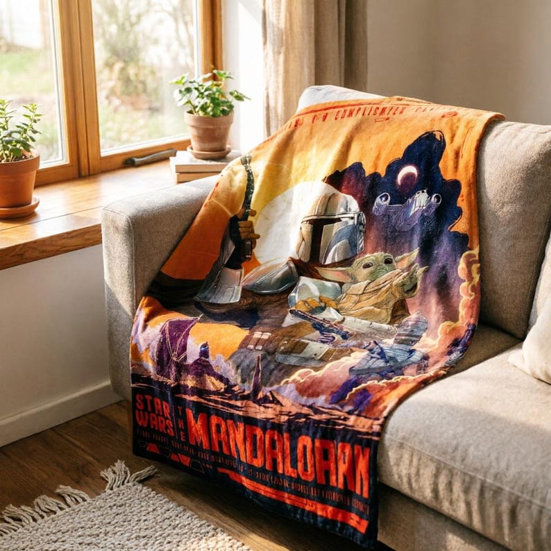 K&ouml;nnte beinhalten: Eine weiche Decke mit einem Star Wars Mandalorian-Design. Die Decke zeigt den Mandalorianer und Grogu vor einem Sonnenuntergangshintergrund, mit den Worten "Star Wars" und "Mandalorian" in fetter Schrift. Die Decke ist &uuml;ber ein graues Sofa drapiert.