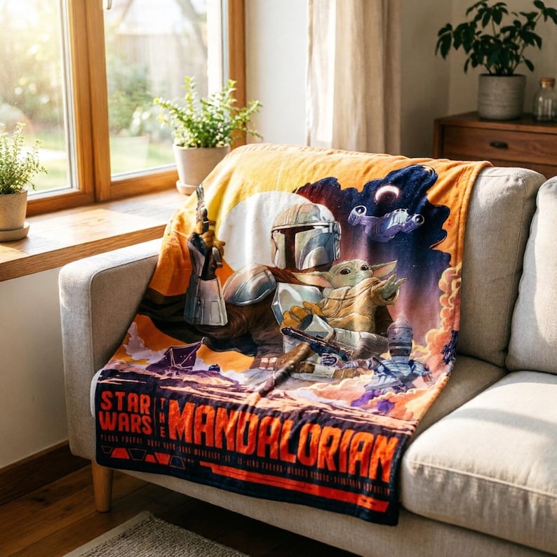 K&ouml;nnte beinhalten: Eine Star Wars: The Mandalorian-Decke mit dem Mandalorianer und Grogu. Die Decke hat ein farbenfrohes Design mit dem Logo der Serie unten. Die Decke ist &uuml;ber ein helles Sofa drapiert.