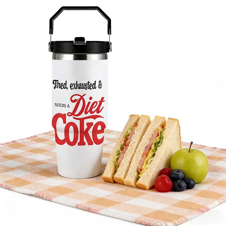 May include: A white insulated tumbler with a black handle and lid, featuring the text "Tired, exhausted & NEEDS A Diet Coke" in black and red. Beside it are two sandwich halves, an apple, and berries on a checkered cloth.