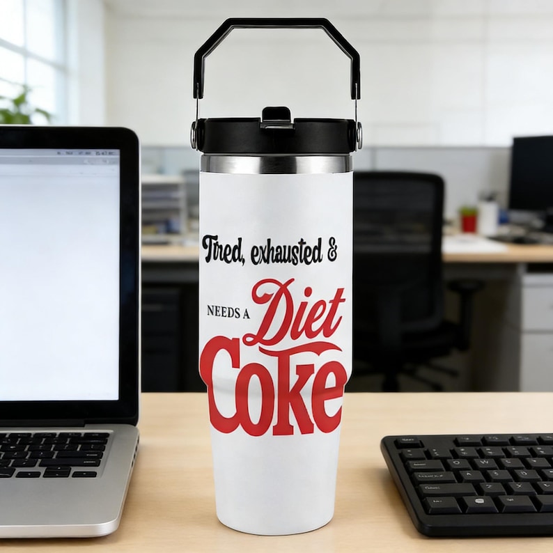 May include: A white insulated tumbler with a black handle and lid. The tumbler has the text "Tired, exhausted & needs a Diet Coke" in black and red script. The tumbler is on a wooden desk.