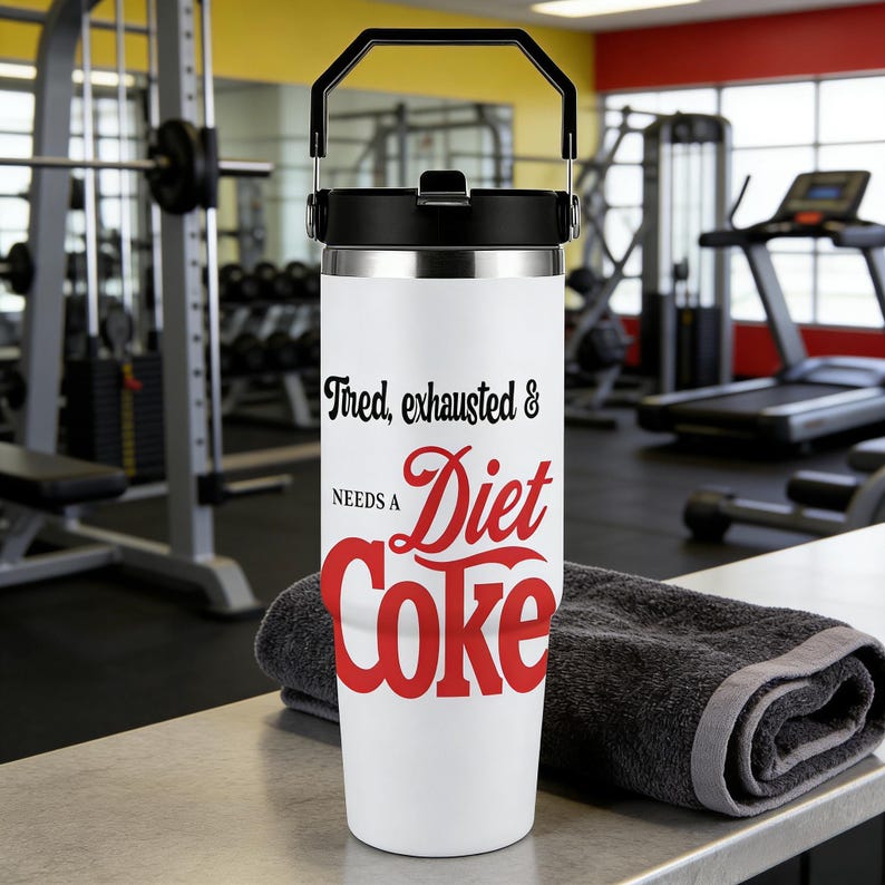 May include: A white insulated tumbler with a black handle and lid. The tumbler features the text "Tired, exhausted & needs a Diet Coke" in black and red. A dark gray towel rests beside the tumbler on a gray surface. The background shows gym equipment.