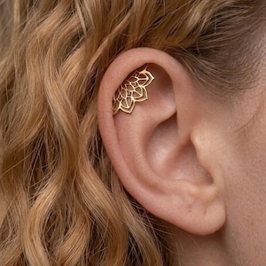 Può includere: Un ear cuff dorato con un delicato design floreale traforato. Il gioiello è indossato sulla parte superiore dell'orecchio, mettendo in risalto gli intricati dettagli del motivo a forma di fiore. Un accessorio elegante.