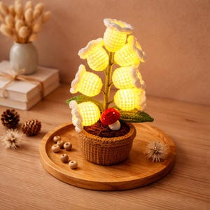 May include: A handcrafted crochet flower lamp with glowing yellow bell-shaped flowers and green leaves, set in a brown crochet pot. A small red and white mushroom sits at the base. The lamp is on a wooden tray.