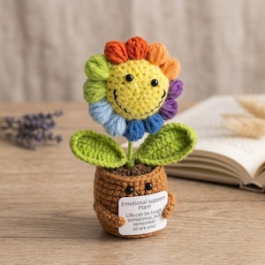 Peut inclure: Une fleur au crochet faite à la main avec un visage souriant et des pétales arc-en-ciel. La fleur est dans un pot marron avec une étiquette indiquant "Emotional support Plant". Le texte sur l'étiquette dit "Life can be tough sometimes, but remember so are you!"