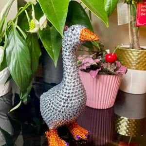May include: A decorative goose figurine covered in silver rhinestones. The goose has an orange beak and feet, with purple accents on the feet. The figurine is displayed on a reflective black surface, with plants in the background.