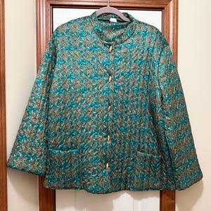 Könnte beinhalten: Türkisfarbene Steppjacke mit floralem Muster in Braun und Gold. Die Jacke hat einen Stehkragen, Holzknöpfe und zwei Vordertaschen. Die Jacke hängt an einem Holzkleiderbügel.