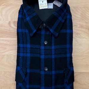 Könnte beinhalten: Gefaltete schwarz-blau karierte Jacke mit Kragen und Knopfverschluss. Die Jacke hat zwei Vordertaschen und ein am Kragen befestigtes Etikett. Die Jacke ist aus einem dicken, warmen Material gefertigt.