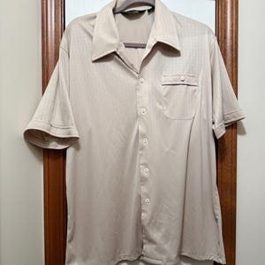 May include: A beige short-sleeved shirt with a button-down front, a chest pocket, and a wide collar. The shirt has vertical stripes and is hanging on a gray hanger. The shirt is made of a shiny material.