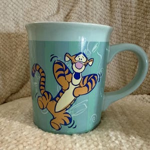 May include: A light blue ceramic mug featuring a graphic illustration of the orange and black striped character Tigger. The mug has a smooth finish, a curved handle, and a vertical striped pattern in shades of blue across the exterior surface.