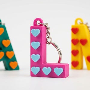 Può includere: Tre portachiavi a forma di lettera colorati: una "A" verde acqua con cuori arancioni, una "L" rosa con cuori azzurri e una "M" gialla con cuori rossi. Ogni lettera ha un portachiavi in metallo.