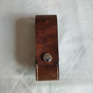 May include: A brown leather case with a silver press stud. The case is rectangular with rounded edges and visible stitching along the sides. The leather has a rich, textured appearance.