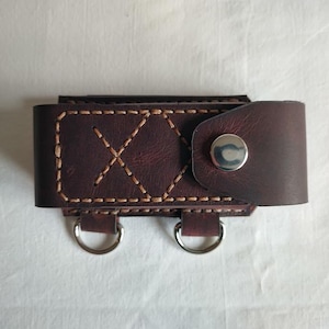 May include: A handcrafted, dark brown leather key holder with a snap closure. The key holder features decorative stitching and two silver-coloured metal rings. The leather has a textured appearance.