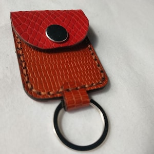 May include: A rectangular keychain pouch featuring a textured orange leather-like material with visible brown stitching along the edges. The top flap is a contrasting bright red textured material secured by a single silver-coloured metal snap button.