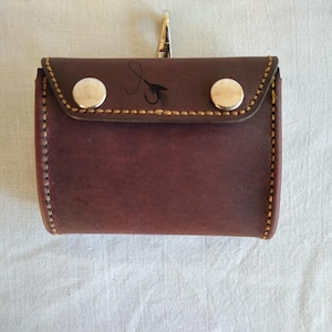 May include: A dark brown leather pouch with a flap closure secured by two silver-toned metal snaps. The flap features a small black graphic of a fishing fly. A silver metal swivel clip is attached to the top centre of the rectangular pouch.