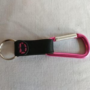 May include: A black leather keychain with a pink stitched "D" and a silver key ring. The keychain is attached to a bright pink carabiner clip with a silver clasp. The carabiner is approximately 7.6 cm long.