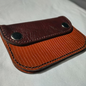 May include: A rectangular wallet featuring a dark brown leather flap with two silver-toned metal snap buttons. The main body of the wallet is constructed from a textured, orange material with a fine vertical ribbed pattern and black stitching.