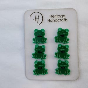May include: A card of six green frog-shaped buttons with black bow ties. The buttons are arranged in two rows of three on a white card with the text "Heritage Handcrafts".