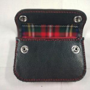 May include: Black leather wallet with red stitching and a tartan interior. The wallet has a snap closure and is open, revealing the tartan fabric. The stitching is visible around the edges.
