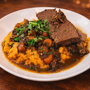 May include: A white plate holds a meal of braised meat and vegetables. The dish features a large portion of cooked meat, sitting atop a bed of mashed sweet potatoes and a medley of carrots and herbs. The food is presented in a restaurant setting.