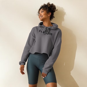 Puede incluir: Una sudadera corta gris con la palabra "Fearless" impresa en negro en el pecho. La sudadera con capucha tiene una capucha con cordón y mangas largas. La modelo lleva unos pantalones cortos de ciclista de color verde azulado.
