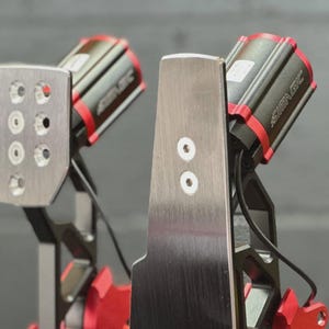 Puede incluir: Un conjunto de pedales para simulador de carreras con acabado en metal cepillado. El pedal izquierdo tiene una cara rectangular con nueve orificios circulares, mientras que el derecho es una placa vertical alta y estrecha con carcasas de motor rojas.