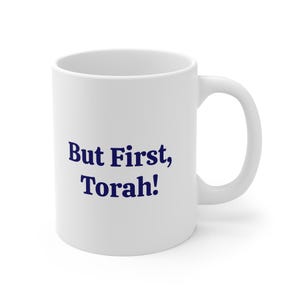 May include: A white ceramic mug featuring the text But First, Torah! printed in dark blue lettering. The mug has a smooth finish, a cylindrical body, and a curved handle on the right side. The design is centred on the front of the white mug.