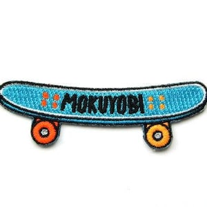 May include: Embroidered patch of a skateboard with the word "MOKUYOBI" in black. The skateboard is light blue with orange and black wheels. The patch has a black border and is on a white background.