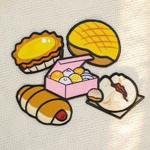 May include: Embroidered patches featuring various baked goods. Includes a yellow tart, a pineapple bun, a pink box of buns, a sausage roll, and a steamed bun with filling. Each patch has a black border.