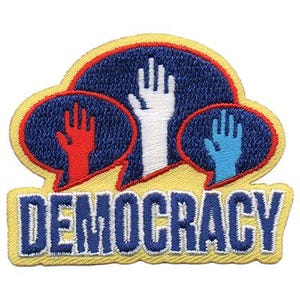 May include: Embroidered patch featuring the word "DEMOCRACY" in blue and white lettering. The patch has a yellow border and includes three raised hands in red, white, and blue, each within a blue speech bubble.