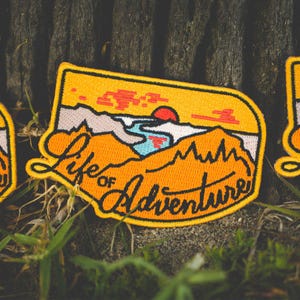 May include: Embroidered patch with a yellow border and a landscape design. The patch features a sunset, mountains, and a river, with the words "Life of Adventure" in orange script.