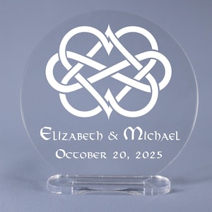 May include: White acrylic wedding cake topper with a Celtic knot design in the shape of two intertwined hearts. The topper is personalized with the names "Elizabeth & Michael" and the date "October 20, 2025".