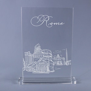 May include: Clear acrylic table sign featuring a white line drawing of Rome's skyline, including the Colosseum and St. Peter's Basilica. The word "Rome" is elegantly scripted at the top. The sign is rectangular with a base.