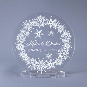 May include: A clear, circular wedding decoration featuring a snowflake design. The names "Kyla & David" and the date "January 24, 2026" are printed in white script. The decoration is mounted on a clear base.