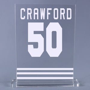 May include: A clear acrylic plaque with a white background and white lettering. The plaque has the name "Crawford" and the number "50" printed on it. There are three white stripes at the bottom of the plaque.