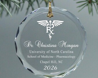 Könnte beinhalten: Ein klarer, facettierter Glasornament mit einer goldfarbenen Aufhängeschlaufe. Der Ornament zeigt ein weißes Caduceus-Symbol und den Text "Dr. Christina Morgan" zusammen mit Universitäts- und Abschlussdetails.