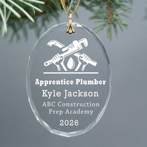 May include: Clear oval-shaped glass ornament with a gold-colored hanging loop. The ornament features a white graphic of crossed plumbing tools and a water droplet. Text reads "Apprentice Plumber Kyle Jackson ABC Construction Prep Academy 2026."