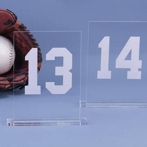 May include: Two clear acrylic signs with white numbers. The sign on the left displays the number 13 and the sign on the right displays the number 14.