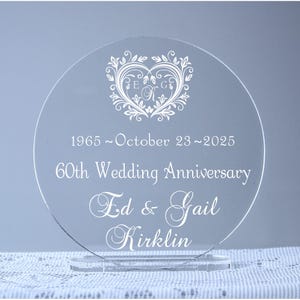 May include: A clear acrylic round plaque with a white engraved design. The design features a heart with a floral pattern and the initials "E & G". The text "1965 ~ October 23 ~ 2025 60th Wedding Anniversary Ed & Gail Kirklin" is also engraved on the plaque.