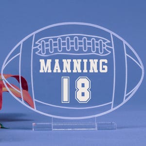 May include: Clear acrylic football-shaped decoration with the name "MANNING" and the number "18" in white block letters. The football design has white outlines and sits on a clear base. The background is a solid blue.