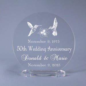 May include: Clear acrylic wedding anniversary keepsake. Features two hummingbird illustrations and a floral design. The text reads "50th Wedding Anniversary" with names and dates. Includes a clear base for display.