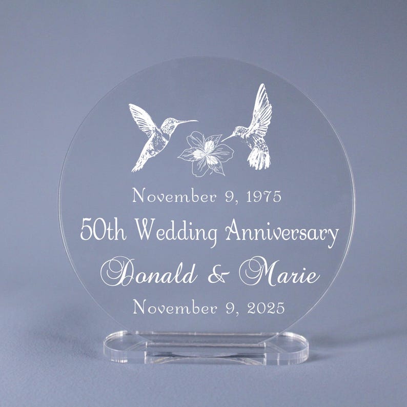 May include: Clear acrylic wedding anniversary keepsake. Features two hummingbird illustrations and a floral design. The text reads "50th Wedding Anniversary" with names and dates. Includes a clear base for display.