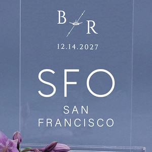 May include: A rectangular clear acrylic sign featuring white printed text. The design includes the letters B and R separated by a small airplane icon, the date 12.14.2027, and the text SFO SAN FRANCISCO. The sign is supported by a clear acrylic base.