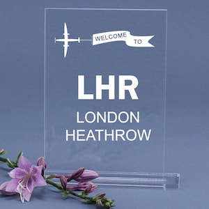 May include: Clear acrylic sign with the text "WELCOME TO LHR LONDON HEATHROW" in white. A white airplane graphic and banner are above the text. A sprig of purple flowers is in the foreground.