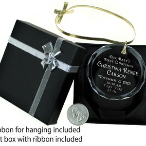 May include: A clear glass Christmas ornament with a gold ribbon. The ornament is engraved with "Our Baby's First Christmas Christina Renee Carson November 6, 2023 10:48 AM 7 LBS 9 OZ 21 IN". The ornament is in a black gift box with a silver ribbon.